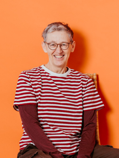 Dor Lötscher sitzt lachend auf einem Stuhl vor einer leuchtend orangen Wand. Dor trägt ein rot-weiss gestreiftes T-Shirt über einem braunen Langarmshirt, eine runde Brille und braune Hose