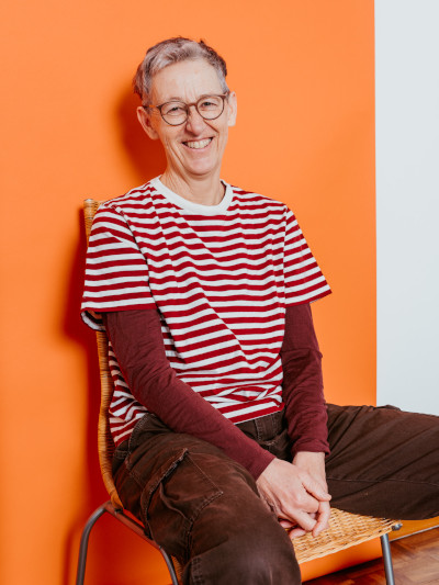 Dor Lötscher sitzt lachend auf einem Stuhl vor einer leuchtend orangen Wand. Dor trägt ein rot-weiss gestreiftes T-Shirt über einem braunen Langarmshirt, eine runde Brille und braune Hose